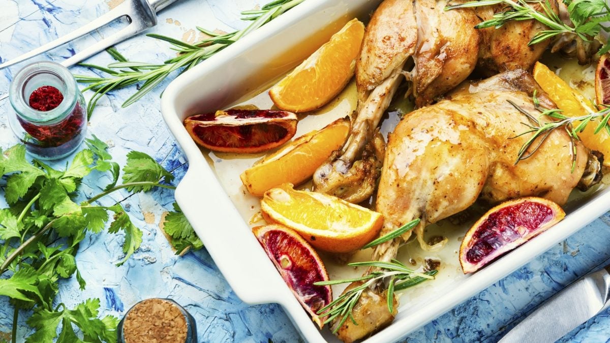 Draufsicht: eine ofenfeste Form mit einem Orangen-Hähnchen sowie Kräutern. Drum herum liegen weitere frische Kräuter sowie Gewürze.