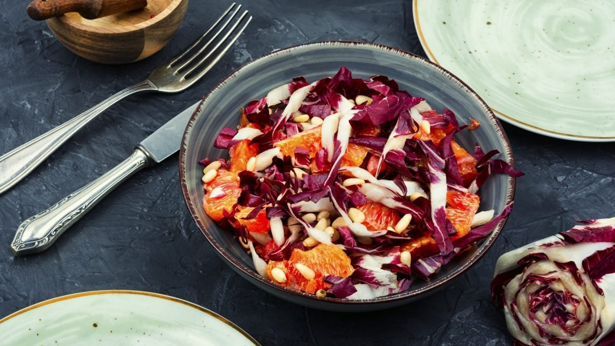Draufsicht: Auf einem dunklen Untergrund steht eine Schüssel Radicchio-Orangen-Salat. Daneben liegen Messer, gabel, ein Radicchio-Strunk sowie zwei Teller, von denen nur die Ränder zu sehen sind.