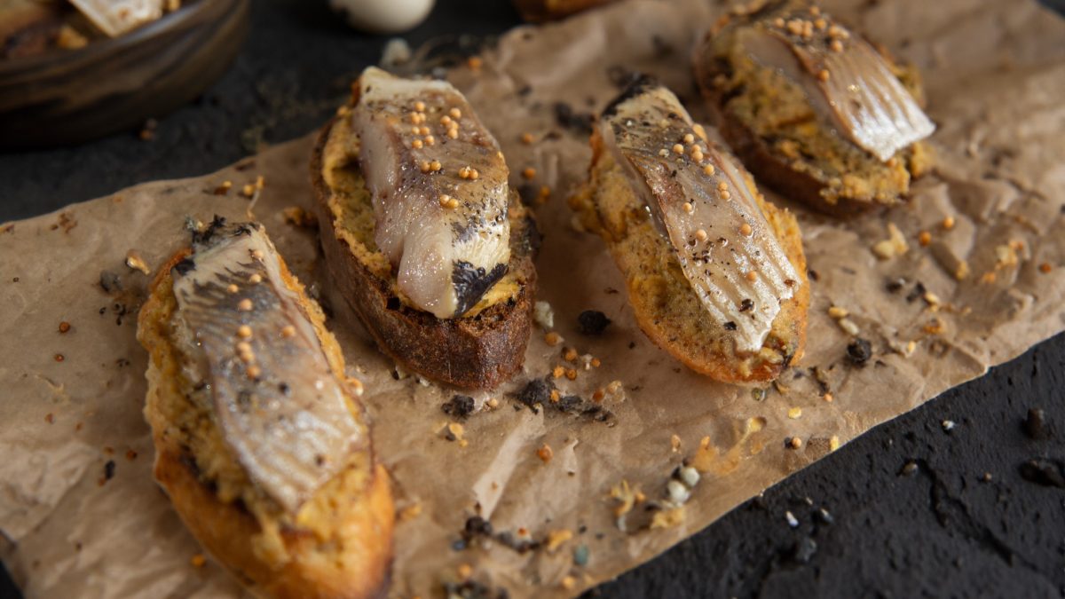 Draufsicht schräg: Ein Backpapier mit Fingerfood. Es handelt sich um mehrere Bruschetta mit Matjes-Hering und eingelegter Senfsaat.