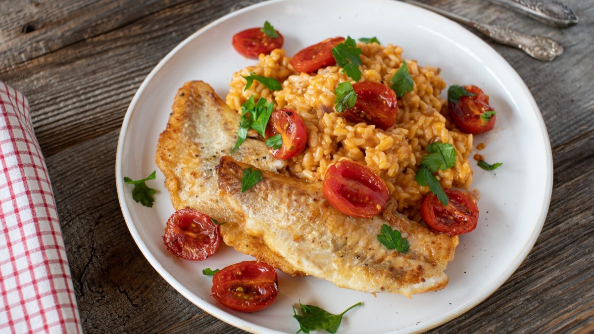 Draufsicht: Auf einem rustikalen Untergrund aus Holz steht ein weißer Teller mit einem Tomaten-Risotto mit Kabeljau. Daneben liegen Besteck und ein kariertes Küchenhandtuch.