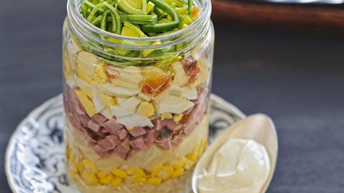 Eiersalat mit Sellerie und Kochschinken im Glas geschichtet auf einem kleinen Teller mit dunklem Untergrund und Löffel.