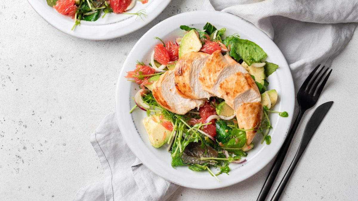 Draufsicht: Ein runder weißer Teller mit einem Hähnchensalat mit Spinat, Avocado und Grapefruit. Daneben liegen Messer und Gabel. Ein weiterer Teller ist teilweise zu sehen.