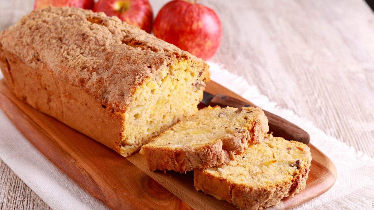 Ein Fantakuchen mit Apfeln auf einem hellen Holztisch mit dunklem Schneidebrett. Alles von der Seite fotografiert.