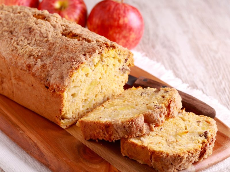 Ein Fantakuchen mit Apfeln auf einem hellen Holztisch mit dunklem Schneidebrett. Alles von der Seite fotografiert.