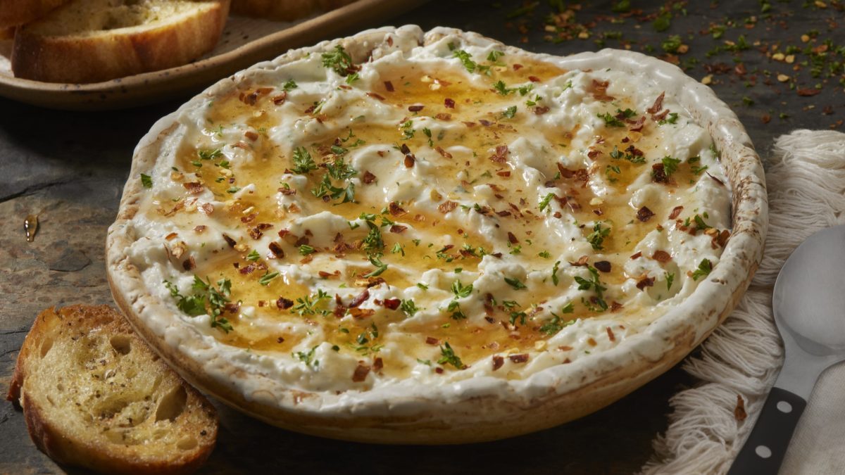 Eine weiße Schale mitd em Feta-Ricotta-Dip auf einem grau-braunem Tisch mit ein paar geröstetn Brotscheiben im Hintergrund. Alles sehr nah von der Seite fotografiert.