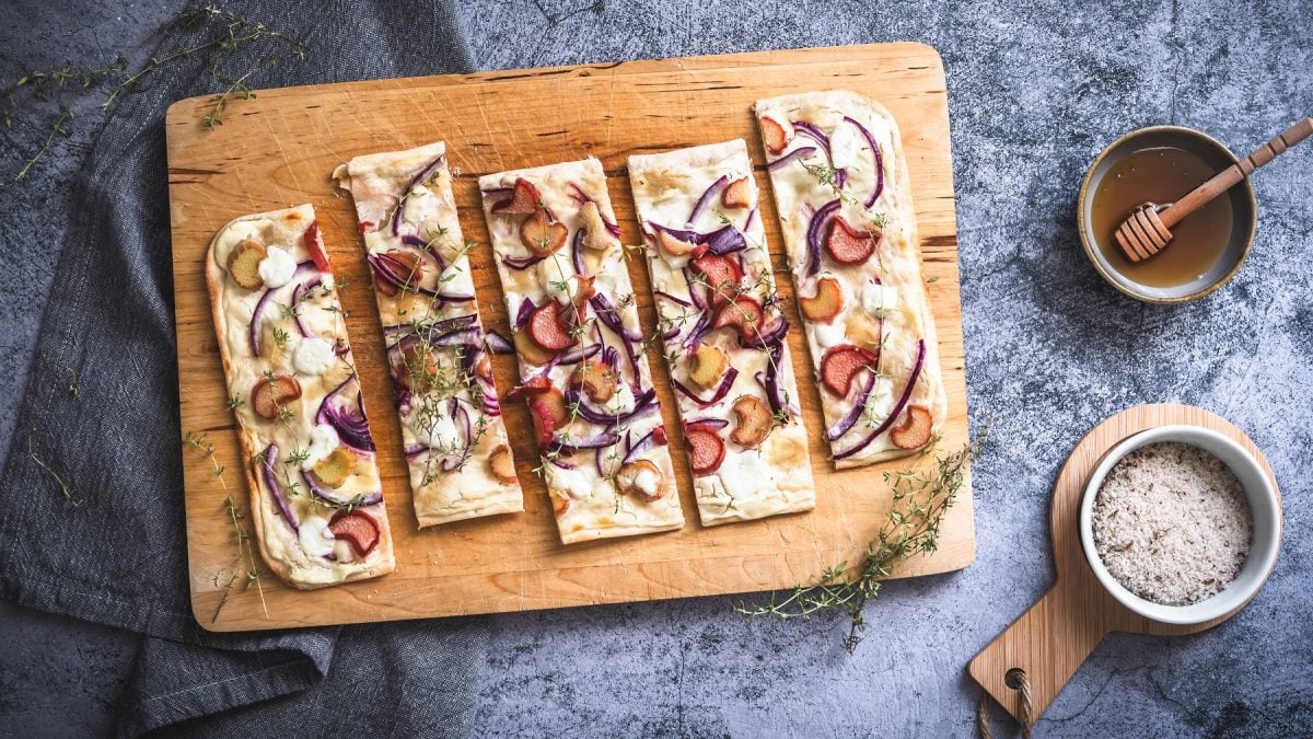Flammkuchen mit Rhabarber, roten Zwiebeln, Thymian und Ziegenfrischkäse auf einem Holbrett. Daneben eine Schale mit Honig und eine Schale mit Meersalz.