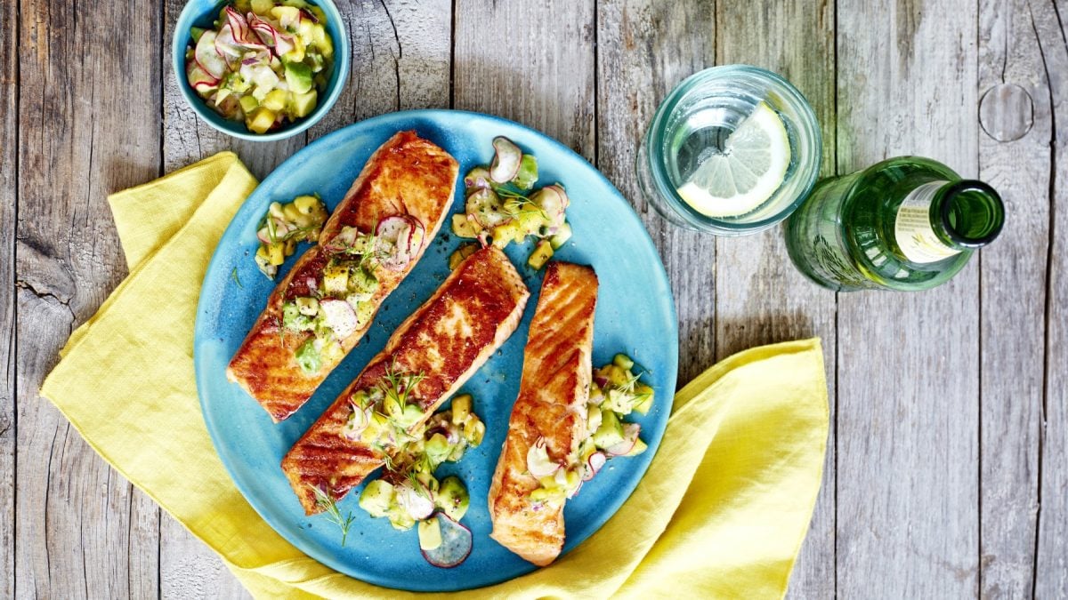 Gewürzlachs mit Radieschen-Avocado-Salsa auf einem blauen Teller auf einem gelben Geschirrtuch. Der Hintergrund ist hölzern. Rechts auf dem Bild eine Wasserflasche und ein Glas Wasser, links auf dem Bild eine kleine Schüssel mit Radieschen-Avocado-Salsa.