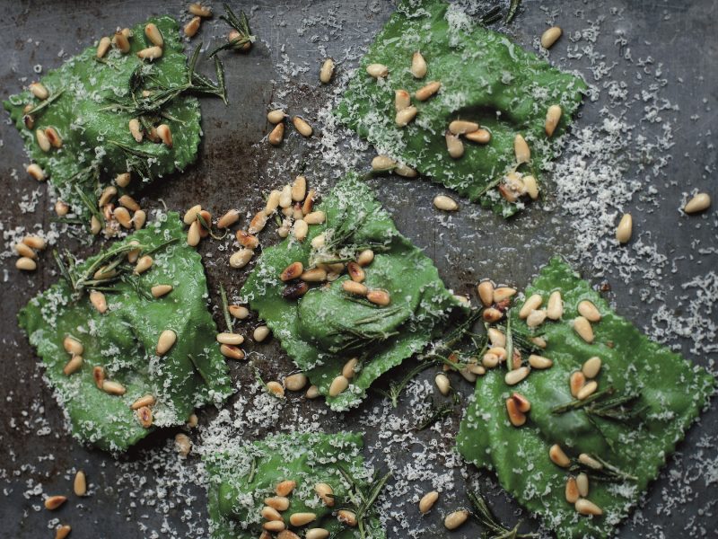 Mehrere grüne Ravioli auf einem grauen Tisch mit Parmesan und Pinienkernen.