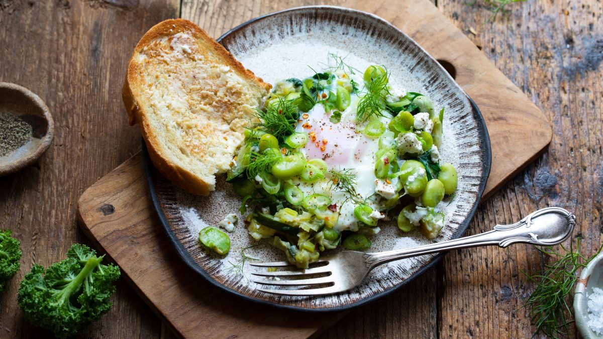 Grünes Shakshuka mit Ackerbohnen, Zucchini, Dill und Spinat auf einem Teller mit gebuttertem Toast.