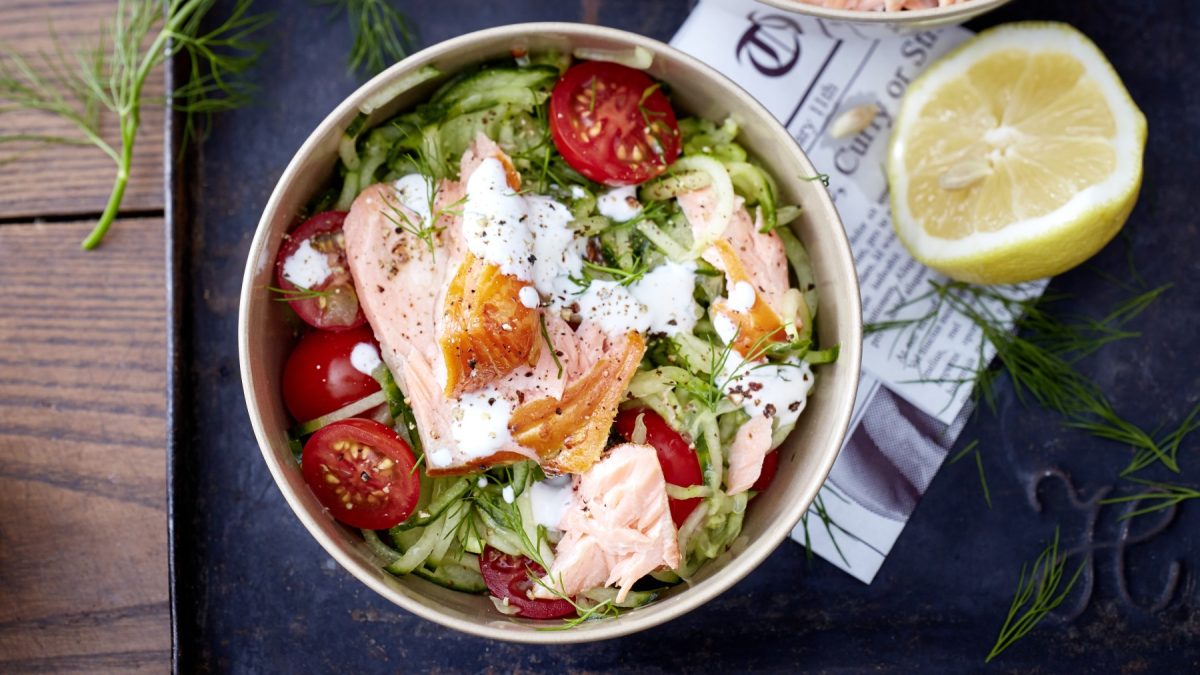 Gurkenspaghetti und Lachs in einem tiefen Teller, daneben eine halbe Zitrone, dunkler Untergrund.