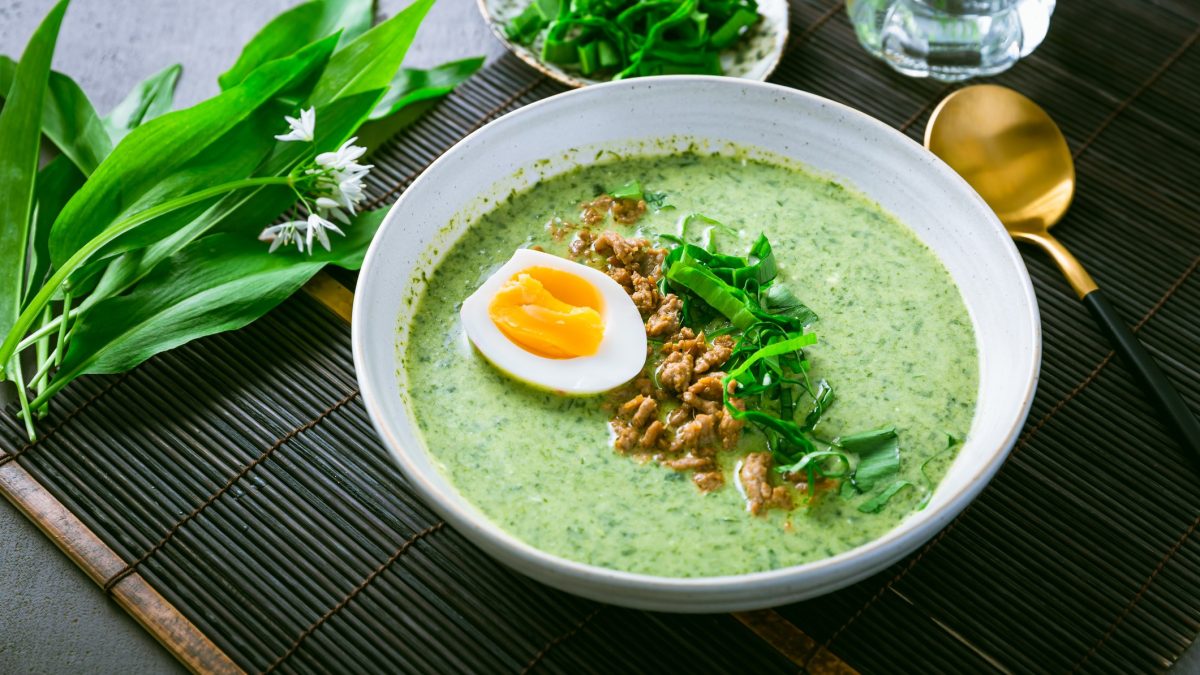 Ein weißer Teller mit der grünen Suppe auf dunklem Untergrund mit frischem Bärlauch und einem goldenen Löffel. Alles von schräg oben fotografiert.