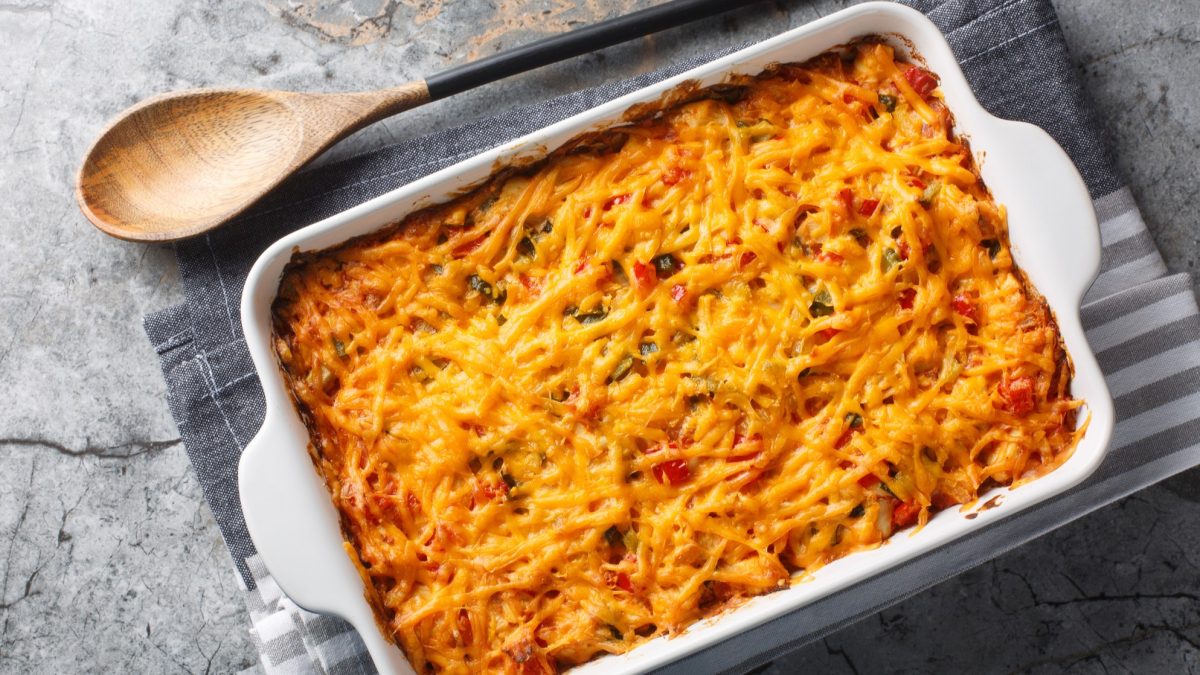 Eine weiße Auflaufform mit dem King Ranch Chicken auf einem Marmortisch mit grauer Tischdecke und Holzlöffel. Alles direkt von oben fotografiert.