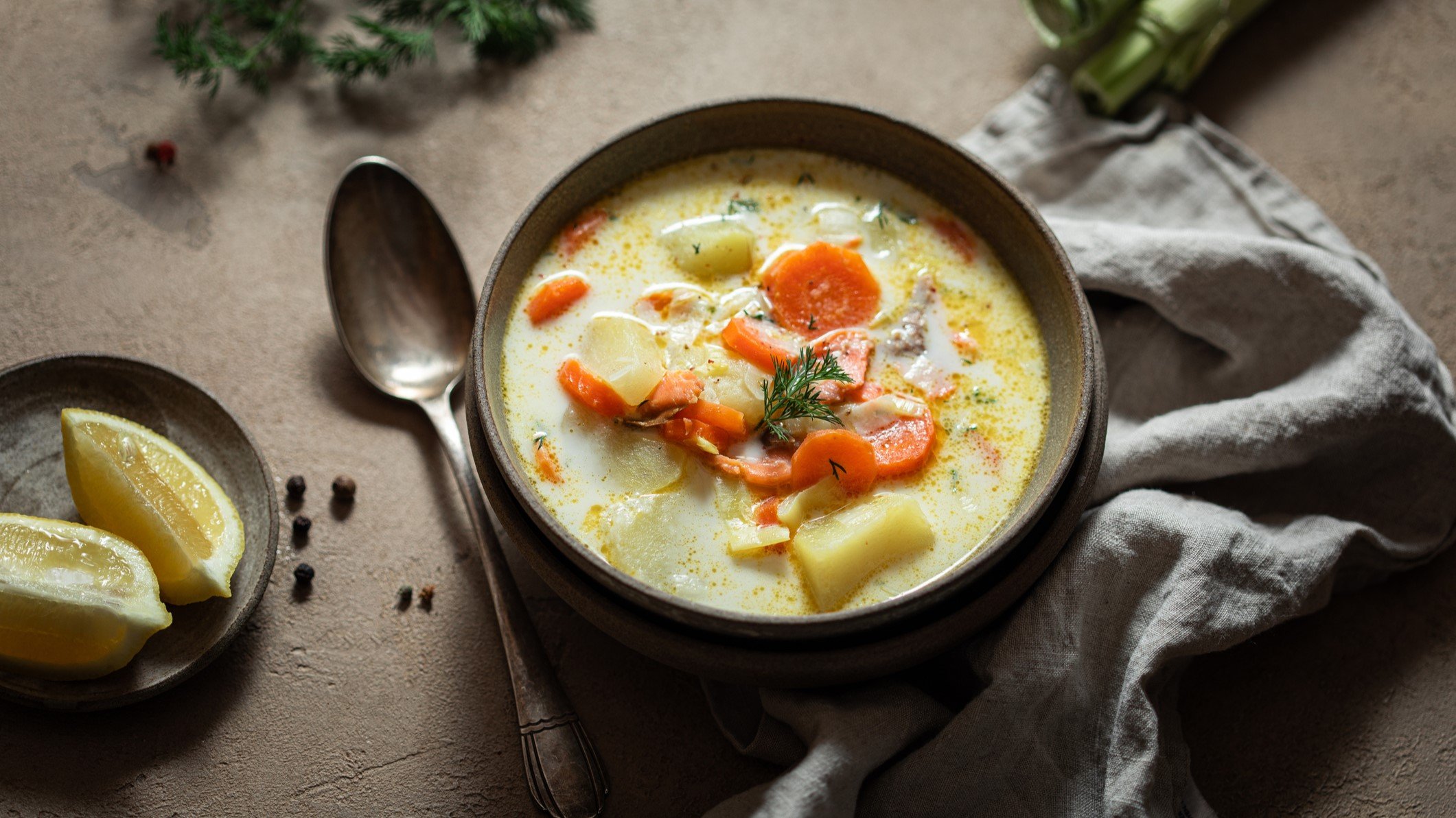 Eine braune Schüssel mit der Lachs-Kartoffel-Suppe auf einem grauen Tisch mit weiteren Gemüsezutaten. Alles von schräg oben fotografiert.