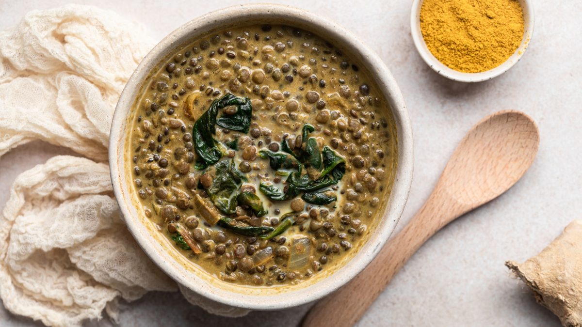 Eine beige Schale mit der Suppe auf einem hellen Tisch, mit Currypulver, Holzkelle und weißem Tuch. Alles direkt von oben fotografiert.