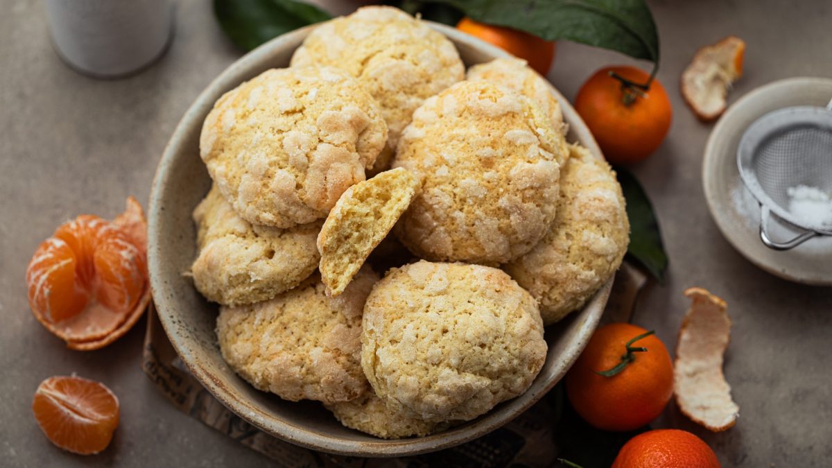 Mehrere Mandarinen-Crinkle-Cookies in einer grauen Schale auf einem grauen Tisch von schräg oben fotografiert. Daneben viele geschälte Mandarinen.