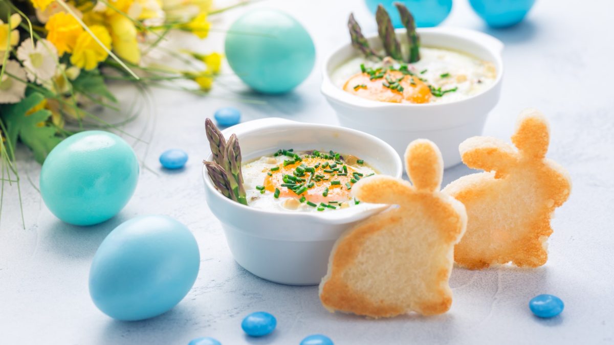 Zwei weiße Schalen mit dem Gericht auf einem hellen Tisch. Daneben viel blaue Osterdeko ein gelber Blumenstrauß und zwei Toastbrote ausgestochen als Osterhase. Alles von der Seite fotografiert.
