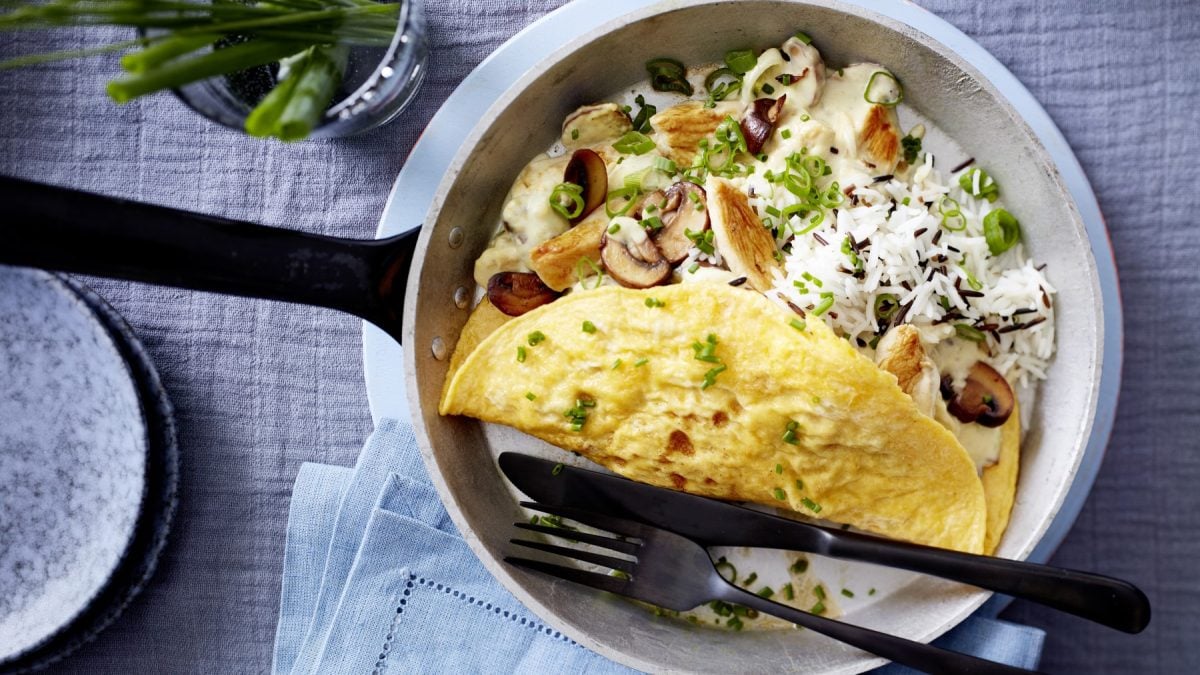 Omelett mit Hähnchen-Pilz-Ragout in einer Pfanne auf einem grauen Hintergrund. Oben auf dem Foto Schnittlauch, unten auf dem Foto ein Geschirrtuch und links ein kleiner Teller.