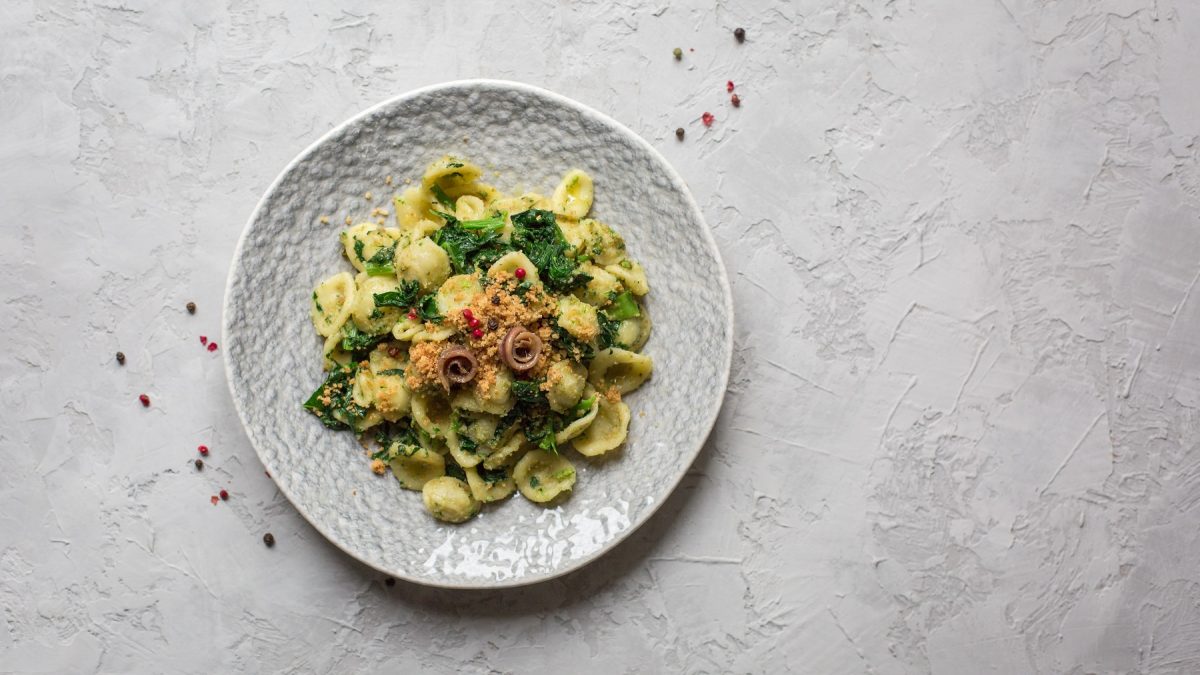 Draufsicht von Orecchiette mit Brokkoli und Sardellen auf einem weißen Teller. Daneben rosa Pfefferkörner.