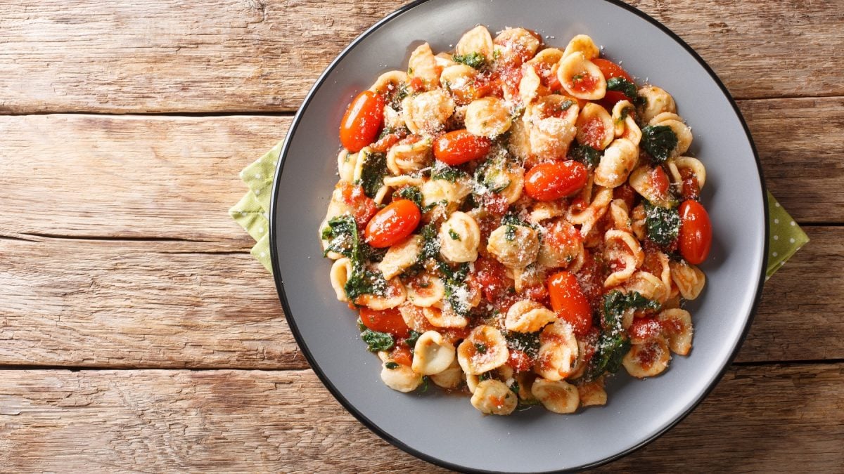 Draufsicht auf Orecchiette mit Spinat und Tomaten auf einem grauen Teller auf einem Holztisch.