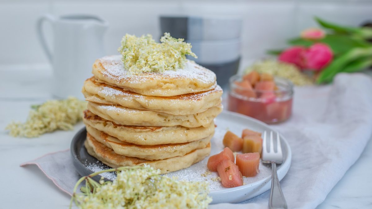 Gestapelte Pancakes und daneben das Rhabarber-Holunderblüten-Kompott. Deko aus frischen Holunderblüten.