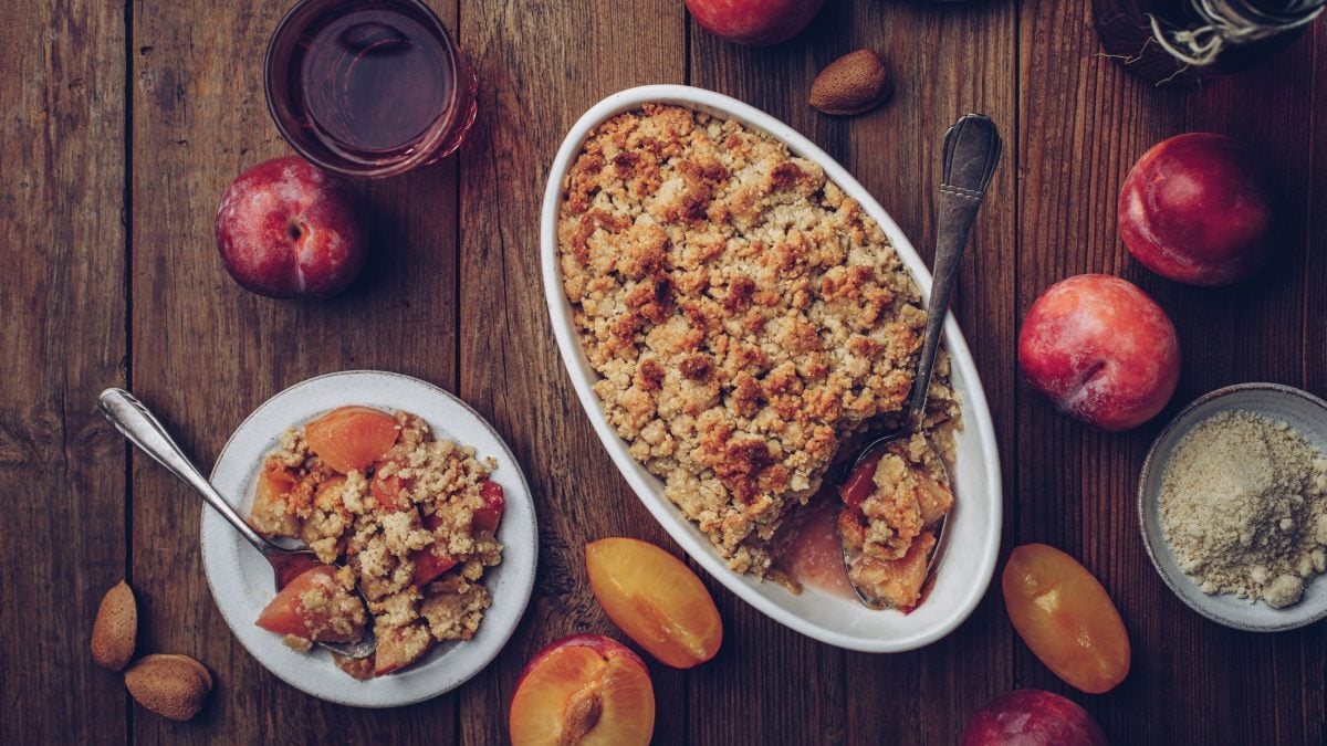 Eine weiße Auflaufform mit dem Pflaumen-Crumble auf einem rustikalen Holztisch mit angeschnittenen Pflaumen. Alles direkt von oben fotografiert.