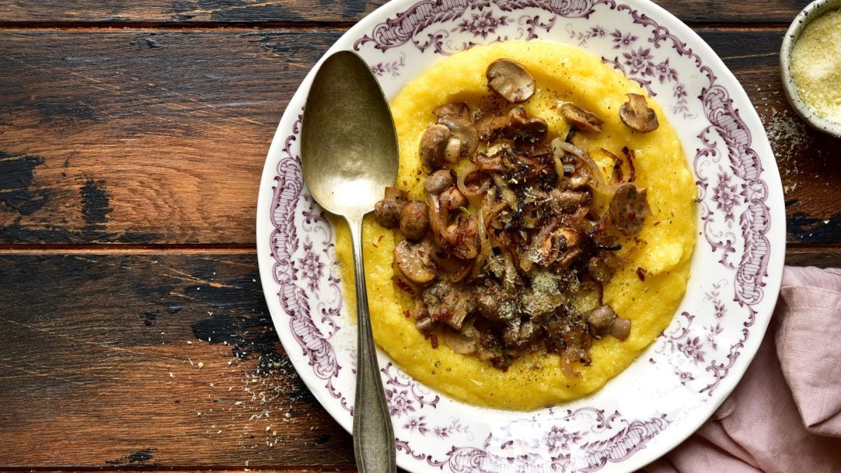 Polenta mit karamellisierten Pilzen in einem Teller. Links ein Löffel. Der Hintergrund ist hölzern.