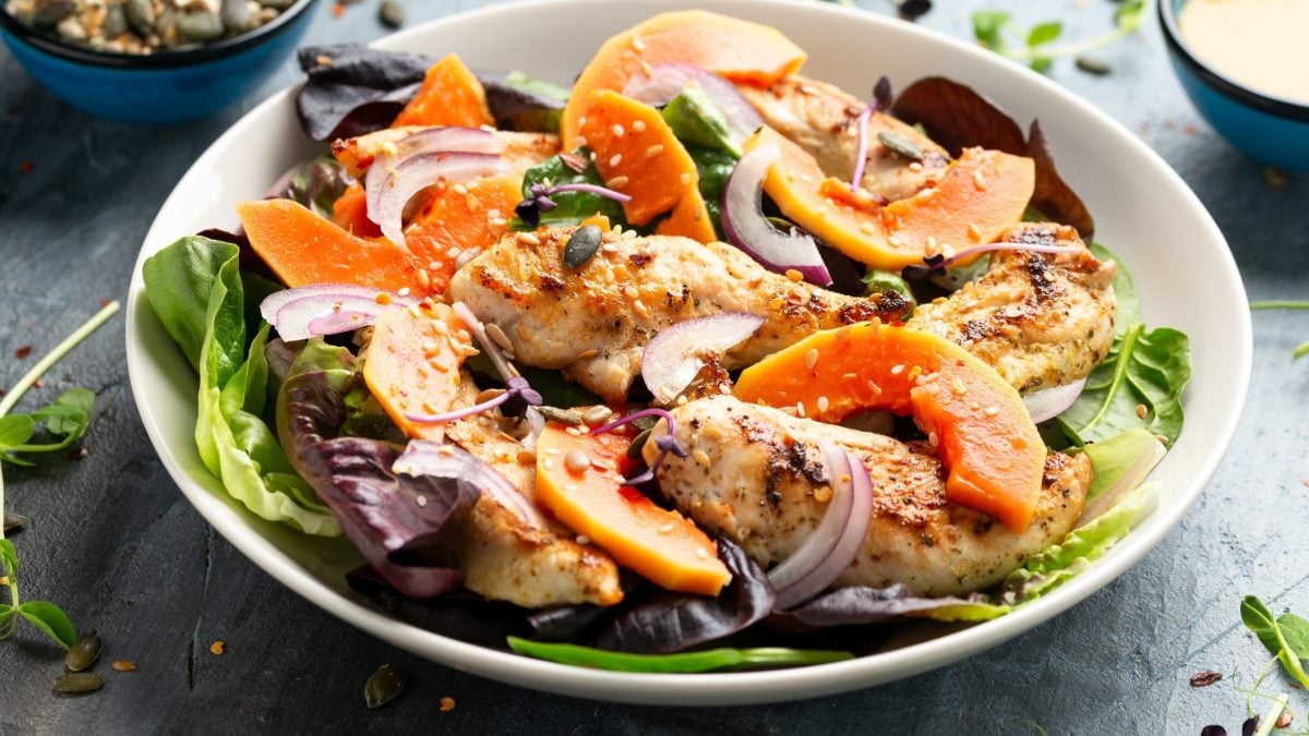 Draufsicht, schräg: ein weißer Teller Putensalat mit Papaya und Sesam sowie Blattsalaten steht auf einem dunkleren Untergrund.