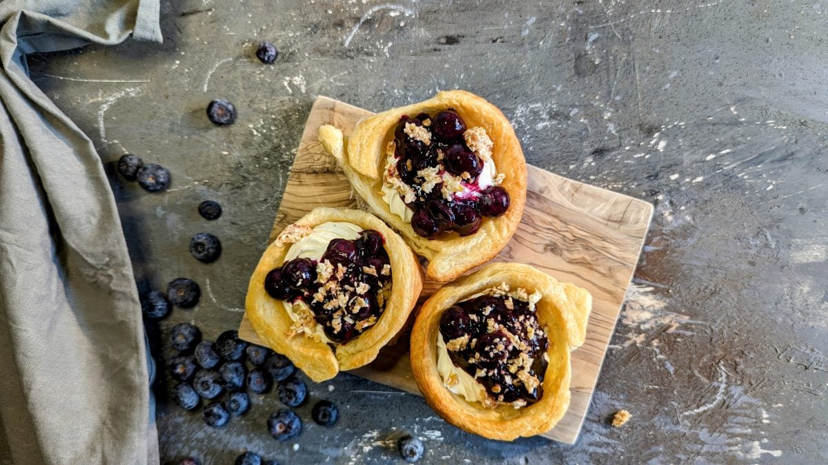 Drei gefülkte Cruffins mit Blaubeerragout auf einem kleinen Holzbrett und einem Steintisch mit Blaubeeren drumherum. Alles direkt von oben fotografiert.