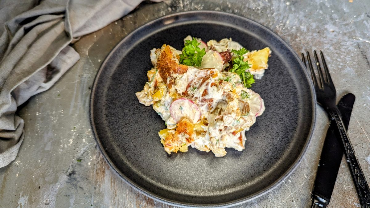 Ein schwarzer Teller mit dem Smashed Potato Salad und schwarzem Besteck sowie grauem Tuch auf einem Steintisch. Alles von schräg oben fotografiert.