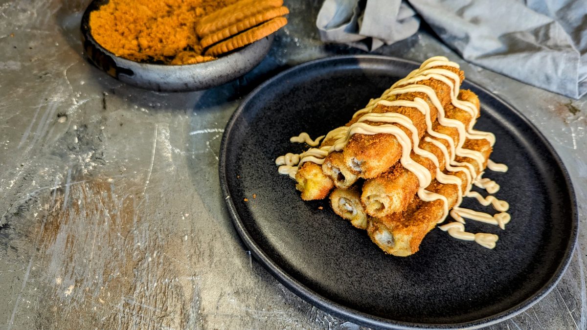 Ein schwarzer Teller mit den Biscoff-French-Toast-Rolls auf einem grauen Tisch mit den Keksen im Hintergrund.
