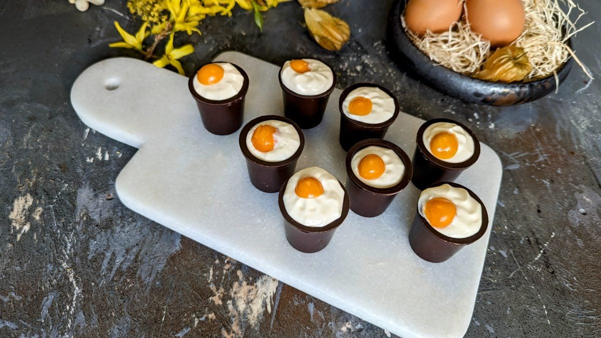 Mehrere Schokobecher mit der Eierlikör-Creme auf einem kleinen weißen Marmorbrett und grauem Tisch. Im Hintergrund Osterdekoration und alles von schräg oben fotografiert.