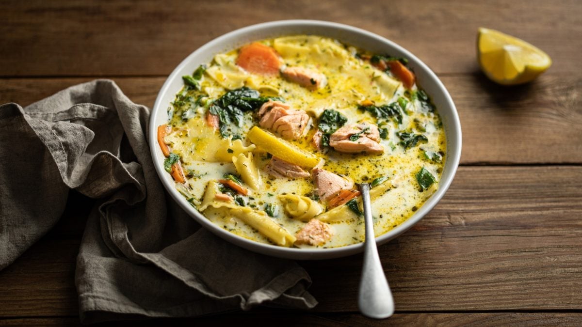Eine helle Schale mit der Suppe auf einem rustikalen Holztisch mit grauem Tuch und Zitronenecke. Alles von schräg oben fotografiert.