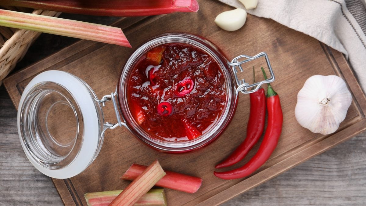 Rhabarber-Chutney in einem Glas. Daneben frische Chilischoten, Knoblauch und Rhabarberstangen.