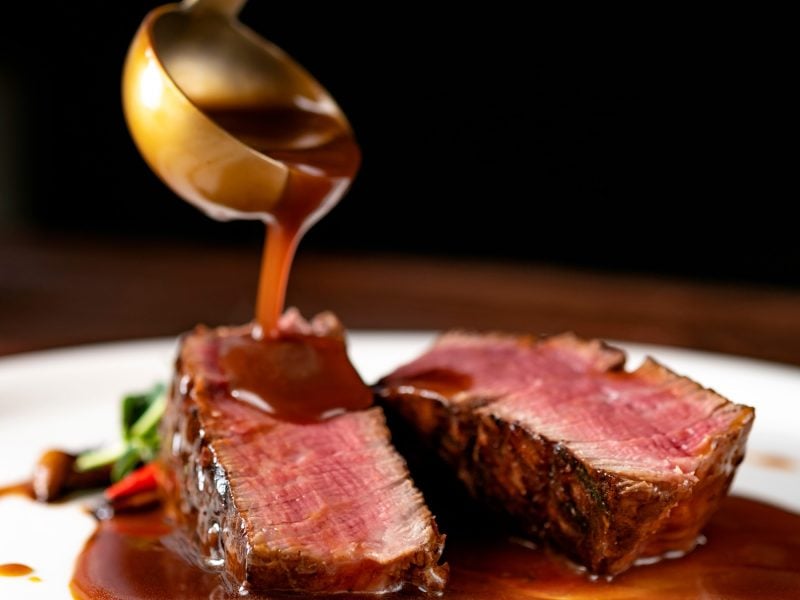 Ein Teller mit Steaks auf den eine braune Sauce gegossen wird.