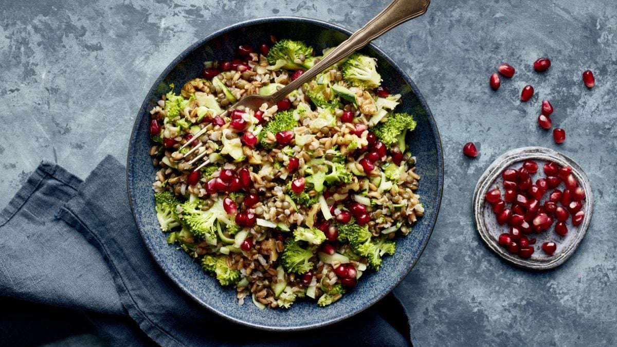 Linsen-Brokkoli-Bowl in einer schwarzen Schüssel auf grauem Hintergrund. Rechts eine kleine Schüssel mit Granatapfelkernen und links unten ein graues Geschirrtuch.