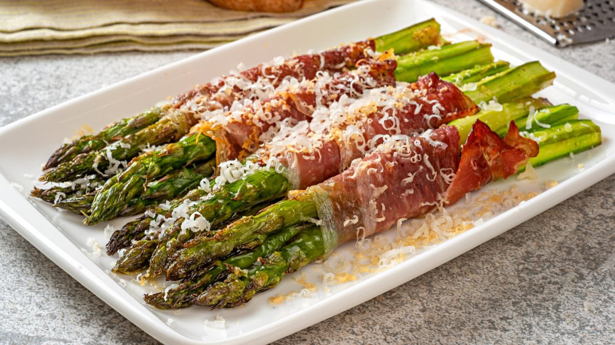 Ein weißer, viereckiger Teller mit den Spargel-Schinken-Päckchen und gehobeltem Parmesan in der Nahaufnahme auf grauen Untergrund. Scharfer Fokus auf die Spargelköpfe.