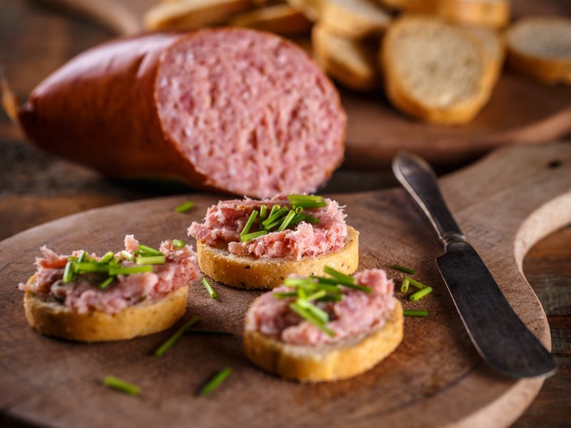 Drei knusprige Baguettescheiben mit fein gestrichener Teewurst und frischem Schnittlauch auf einem rustikalen Holzbrett.