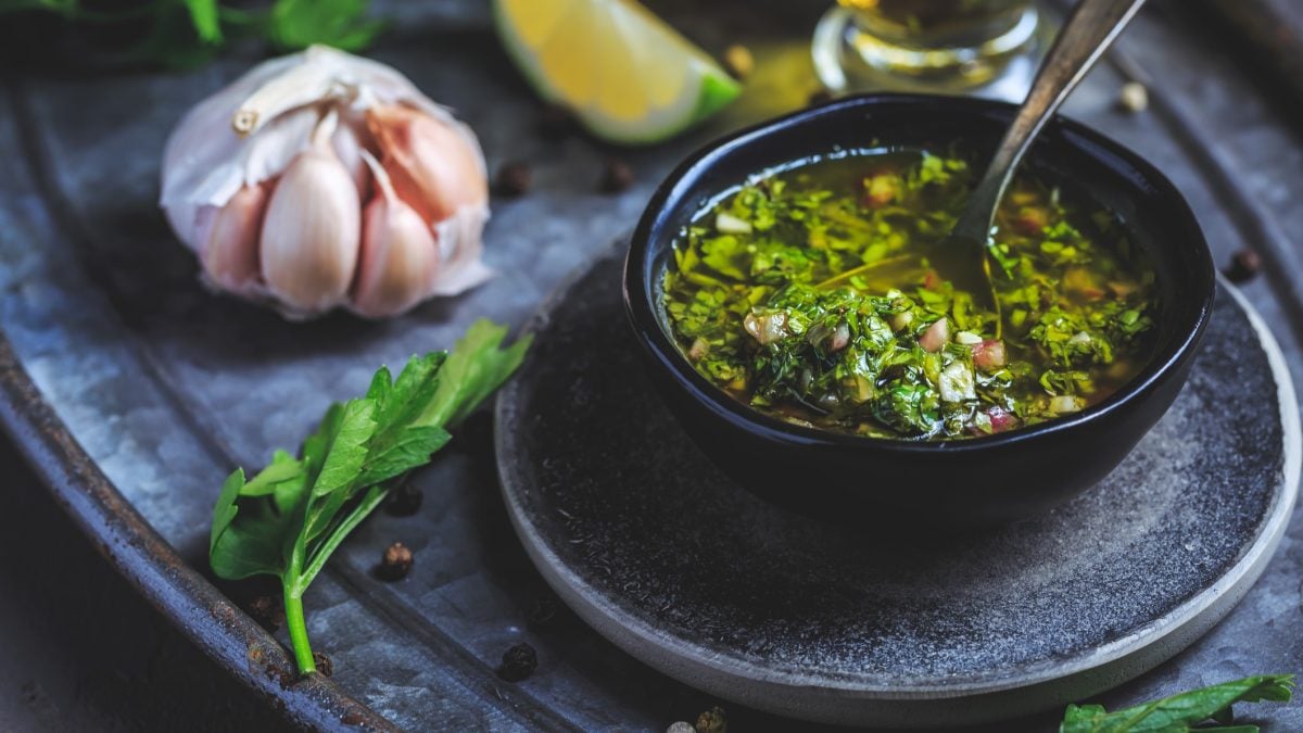 Ein schwarzes Schälchen mit dem Bärlauch-Chimichurri auf dunkelblauem Untergrund, mit Kräutern und Knoblauch. Alles von schräg oben und recht nahe fotografiert.