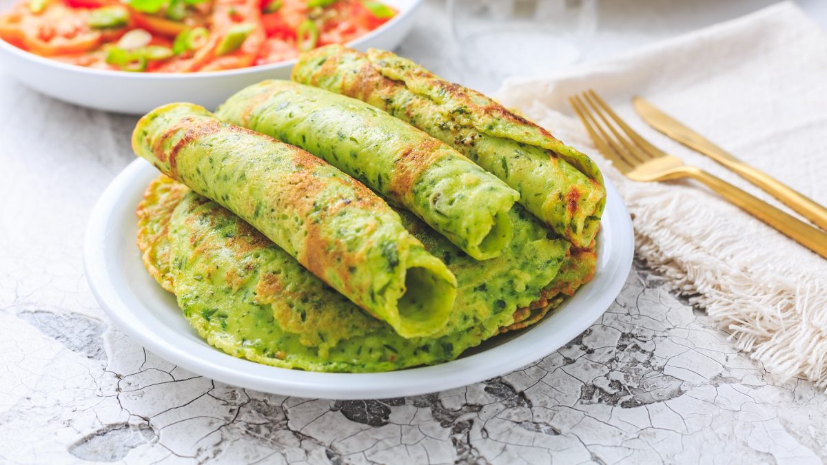 Mehrere Bärlauch-Pfannkuchenröllchen auf einem weißen Teller und hellem Marmoruntergrund. Im Hintergrund eine weiße Schale mit Tomatensalat erkennbar.
