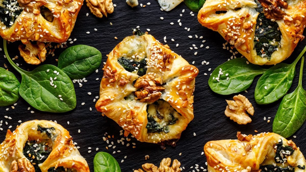 Blätterteigtaschen mit Spinat und Gorgonzola, getoppt mit Walnüssen und Sesam auf dunklem Untergrund.