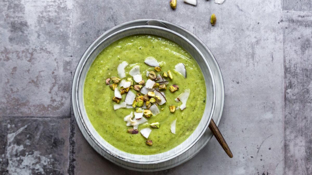 Eine helle Schüssel mit der grünen Suppe auf grauem Untergrund. Alles direkt von oben fotografiert.