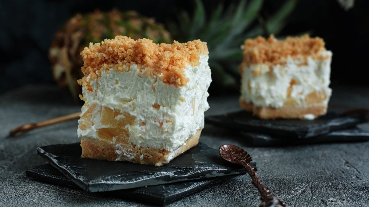 Zwei Stücke mit dem Kuchen auf schwarzen Schieferplatten und dunklem Tisch. Im Hintergrund unscharf eine Ananas und dunkler Hintergrund zu erkennen.