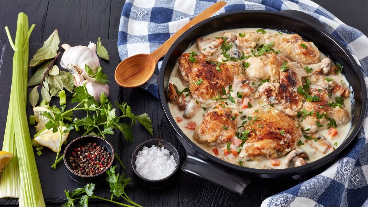 Draufsicht: Eine runder, gusseiserner Topf Coq au Riesling liegt auf einem dunklem Untergrund. Drum herum liegen Zutaten sowie ein blau-weiß kariertes Küchentuch.
