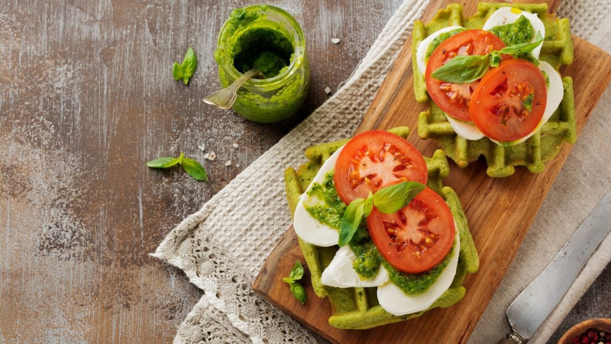 Draufblich: Ein Holzbrettchen mit zwei Caprese-Waffeln, die mit Mozzarella, Basilikum, Pesto und frische Tomaten belegt sind.