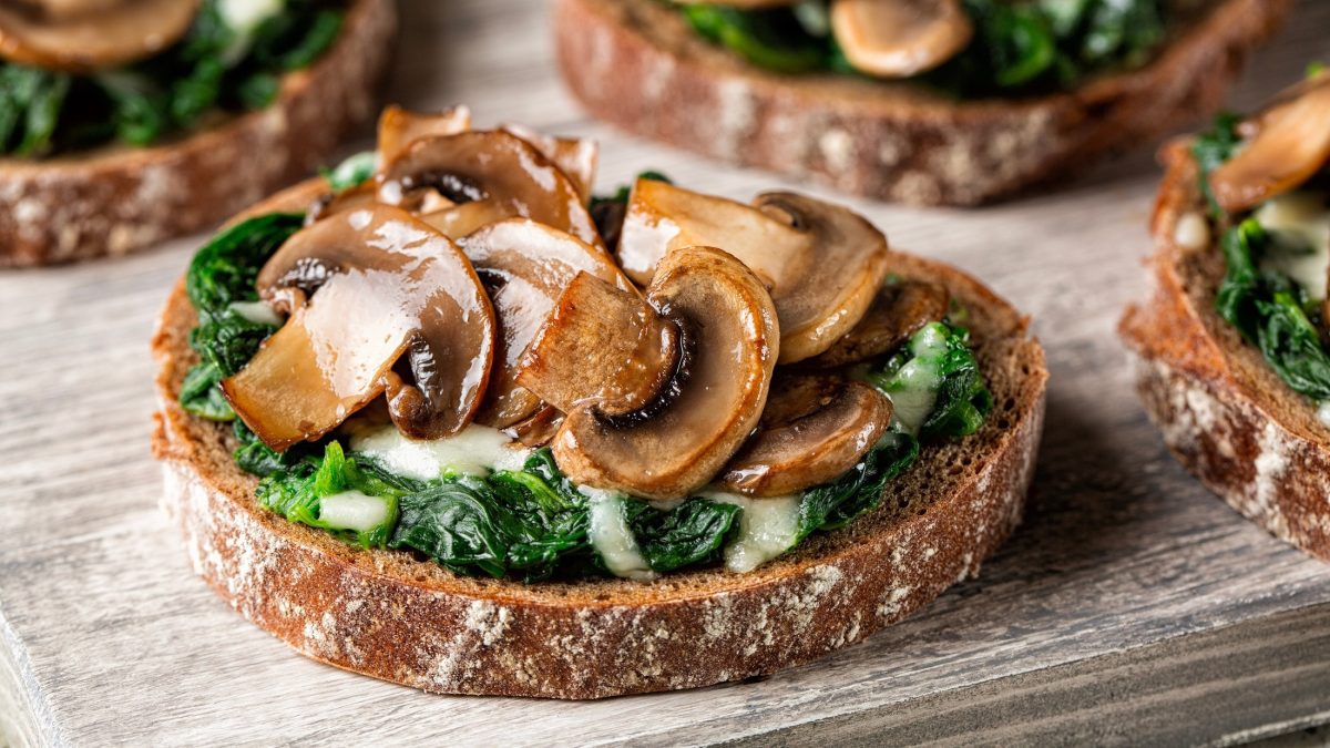 Draufsicht, schräg: ein Cheese Sandwich mit Spinat und Champignons aus der Pfanne liegt auf einem hellen Holzbrett. Im Hintergrund sind drei weitere Brote teilweise zu sehen.