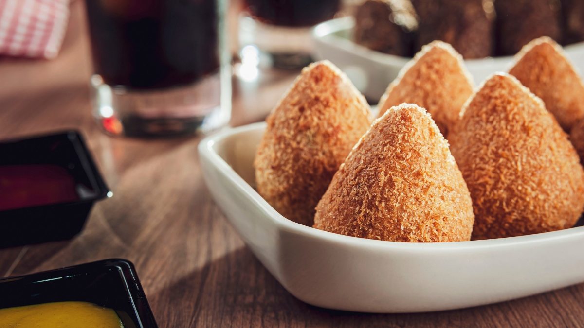 Frontal: Fümf Coxinha de Frango stehen in einer weißen , flachen Schale auf einem Tisch. Es handelt sich um brasilianische, frittierte Snacks aus Hühnchenfleisch.