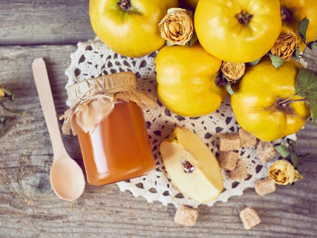 Draufsicht: frische Quitten die man vielleicht roh essen kann, ein Glas Quittengelee sowie ein Löffel aus Holz.