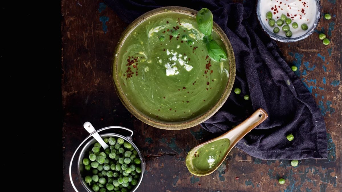 Draufsicht. Eine runde Schüssel Erbsensuppe mit Feta auf einem dunklem Untergrund. Daneben sind eine kleine Schale mit TK-Erbsen, ein Löffel und eine Schale mit einer weißen Creme.