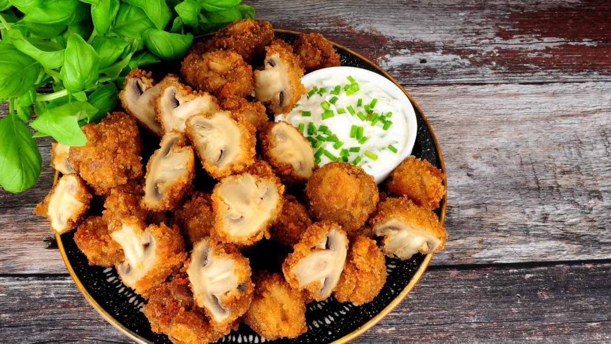 Draufsicht: Eine Teller frittierte Champignons mit einem schälchen weißer Sauce. An einem Rand sind frische Basilikum-Blätter.
