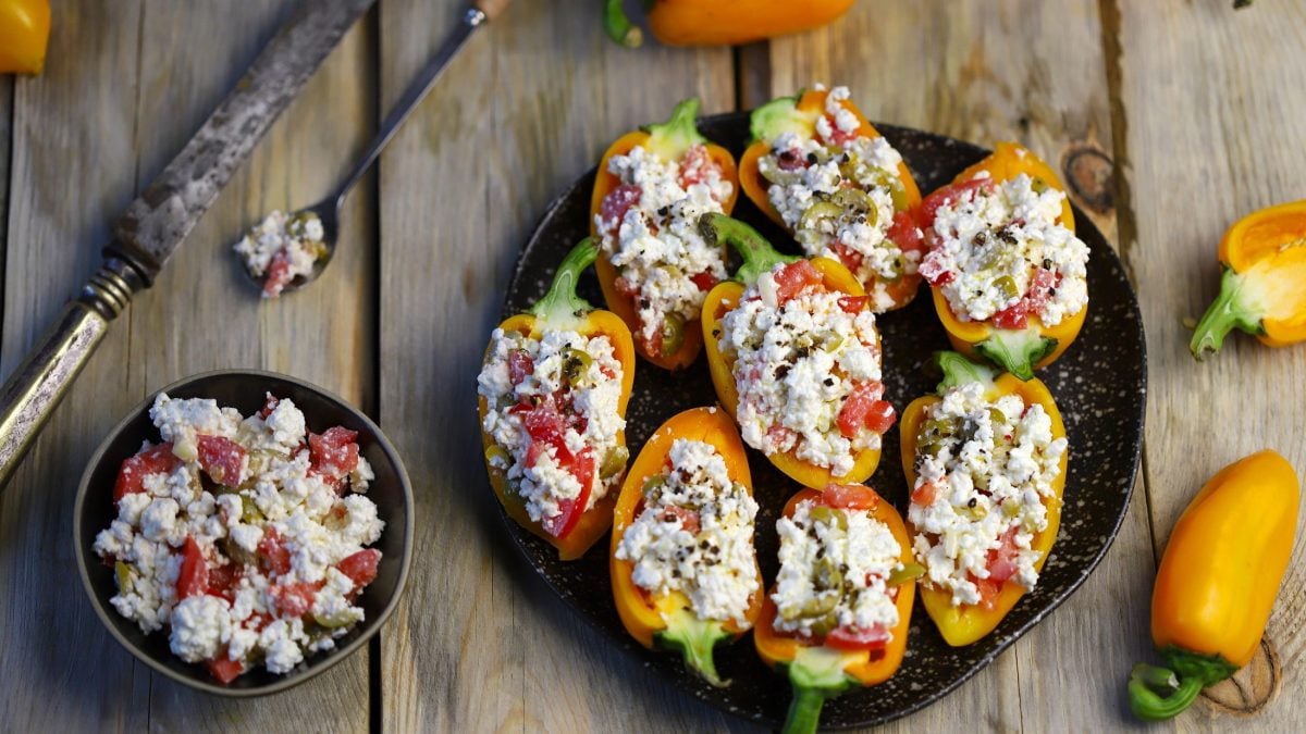Draufsicht: Auf einem Untergrund aus Holz liegt ein rundes Tablett. Auf dem sind mehrere gefüllte Mini-Paprika. Daneben liegen intakte gelbe Paprikaschoten.